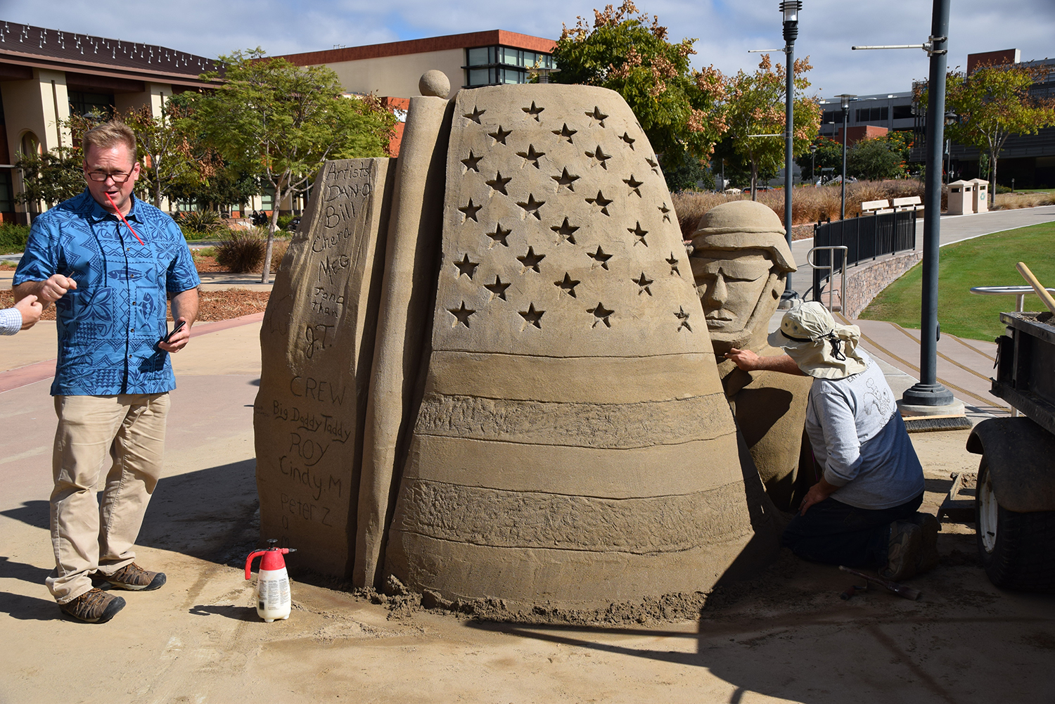 Dan Gutowski left directs the construction of a sand sculpture for veterans day. The sculpture is of a soldier's head with a helmet and a sand wall with stars carved into it.
