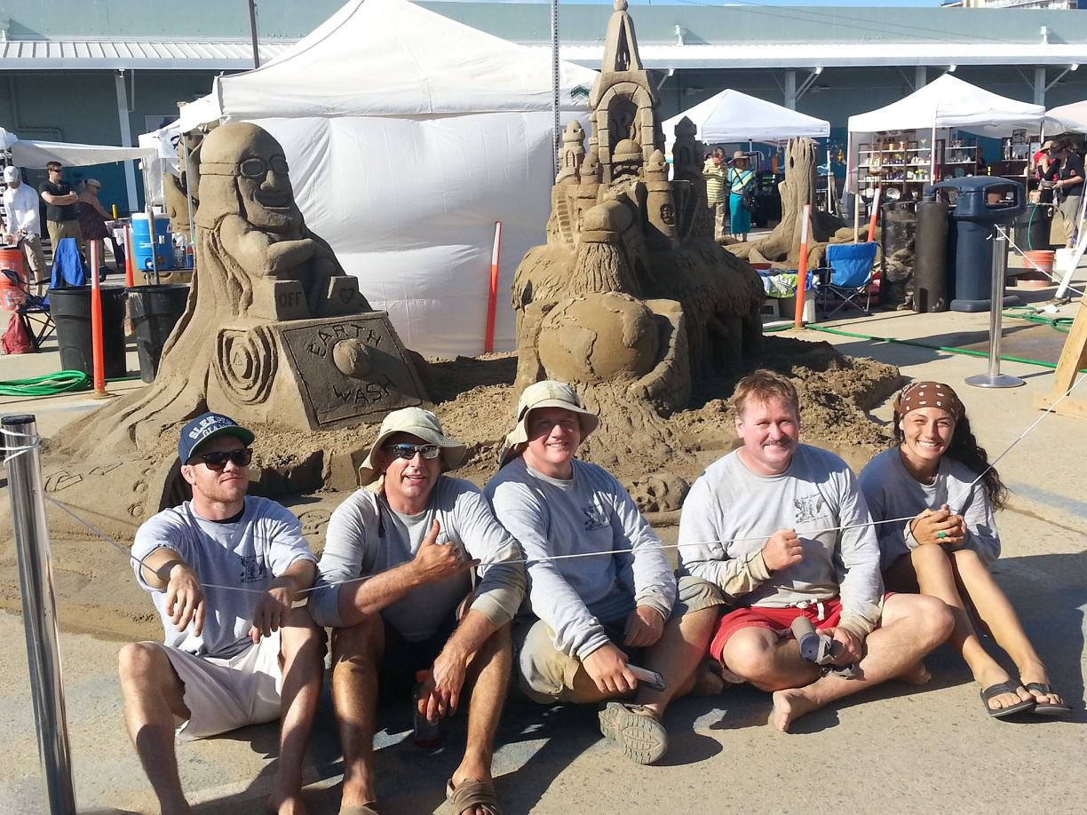 5 people at a fair sit in front of 2 sand sculptures. One show a man on a throne with a helmet and a cape and the other is a sand castle on top of a globe.