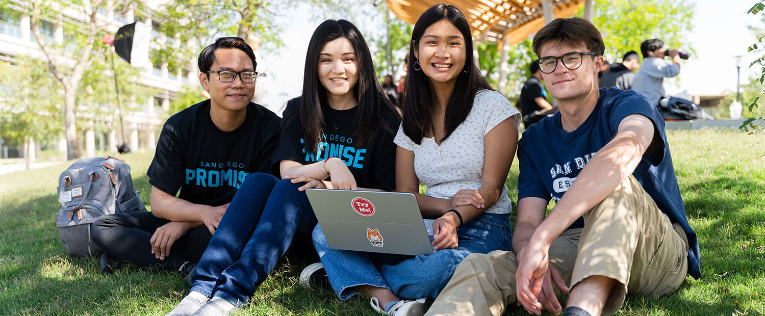 Four students sit on the grass at the Mesa College quad. One is using a laptop. Two of the students are wearing black T shirts that say San Diego Promise in teal.