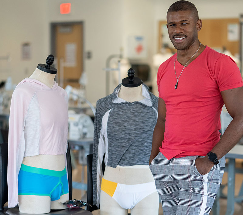 Darius Stokes in a red T shirt and black and white pants stands next to mannequins wearing a gray sweatshirt and a pink sweatshirt that he designed.