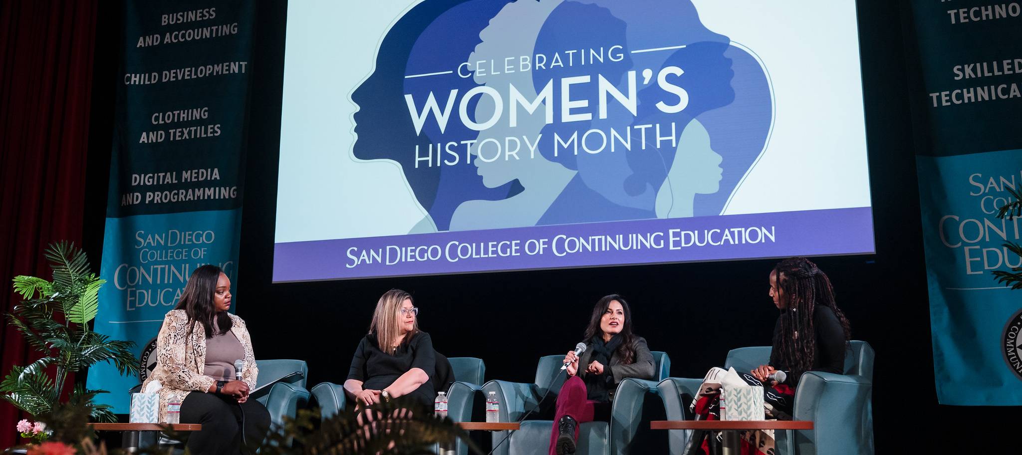 Four women hold a discussion on a stage a large slide is illuminated behind them. The slide shows a purple profile of three women text reads womens history month