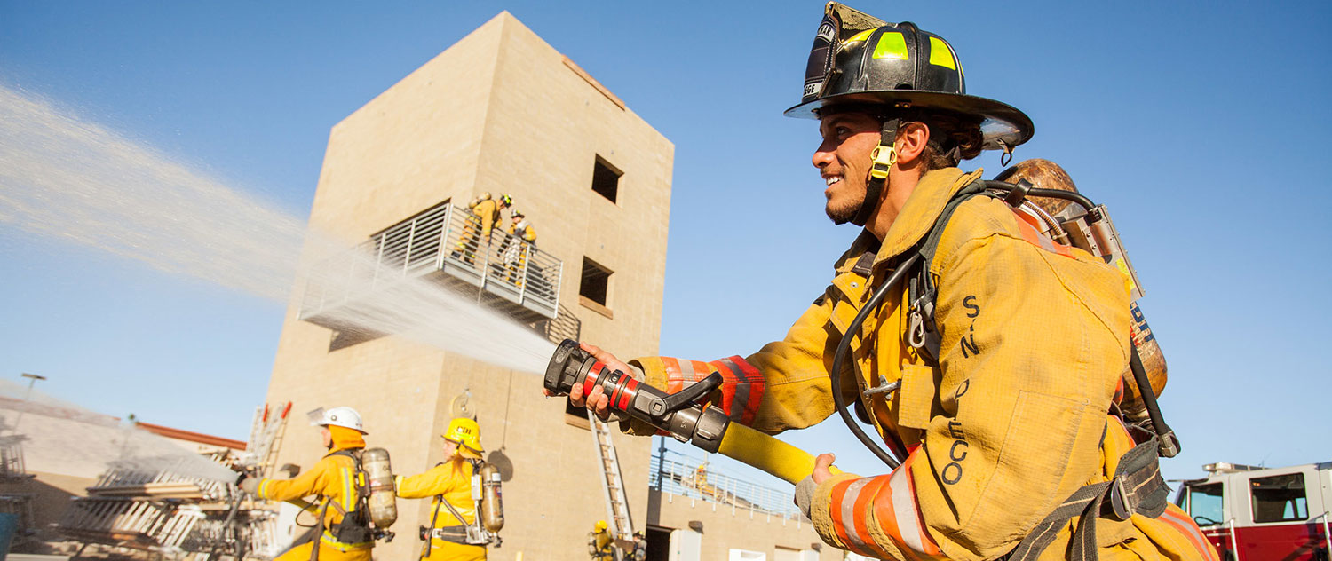 A fire academy student in yellow fire gear uses a fire hose at the training site at Miramar College