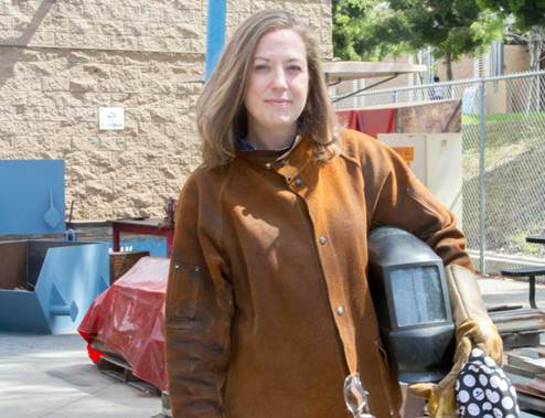 Andrea Cook wears a tan coat welding gloves and holds a welding helmet