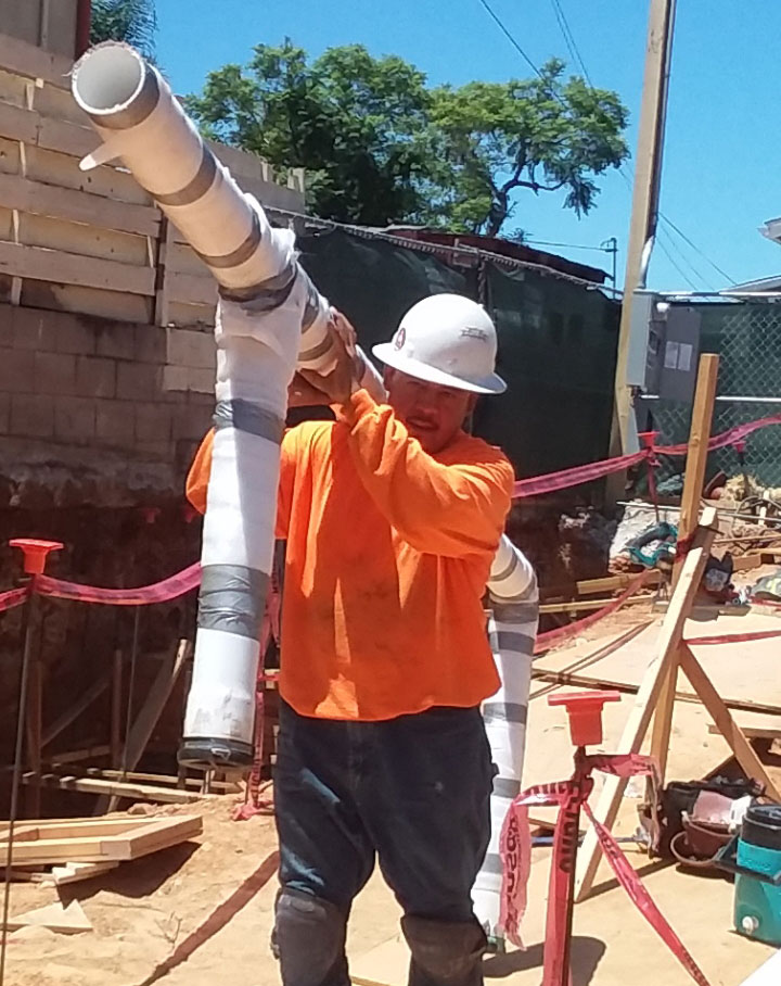 Manuel Cortez carries plastic pipes while wearing and orange shirt and white hard hat on a construction site