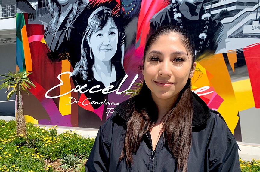 Jeannette Mayo Gallegos wears a black graduation cap and gown and is standing in front of a colorful mural at City College