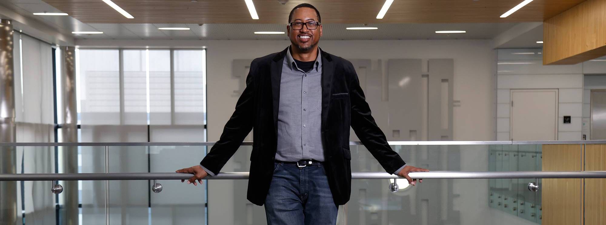 Alphonso Brown wears blue jeans, a blue button down shirt and a black blazer. He is leaning on the railing on the second floor of the North City Campus.