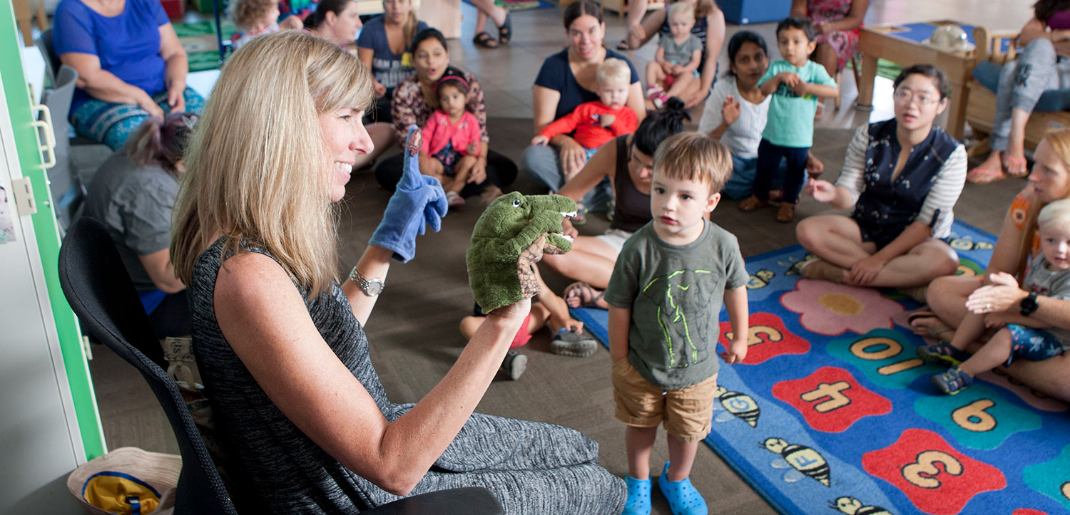 Children and parents sit on the floor and watch an instructor perform a puppet show.