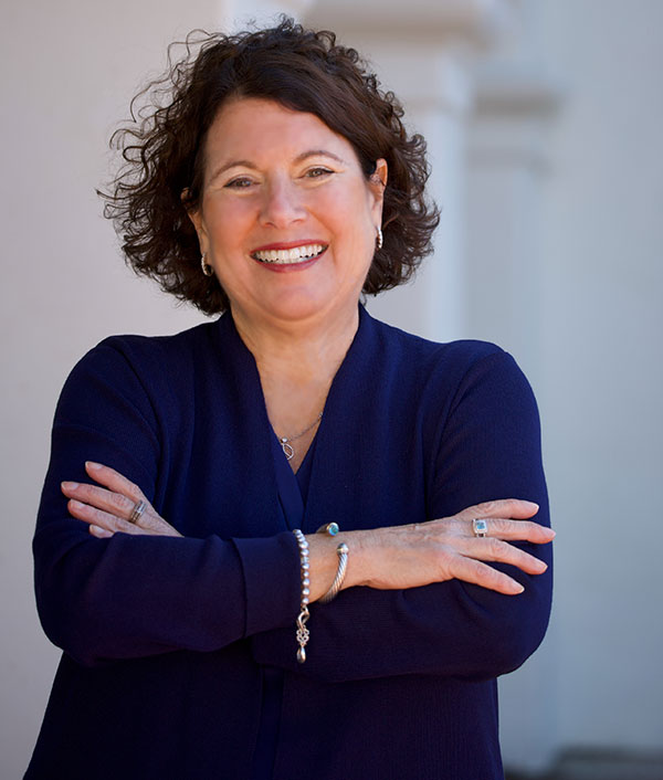 Laurie Coskey portrait photo. She is standing in front of while pillars and wearing a Royal Blue top.
