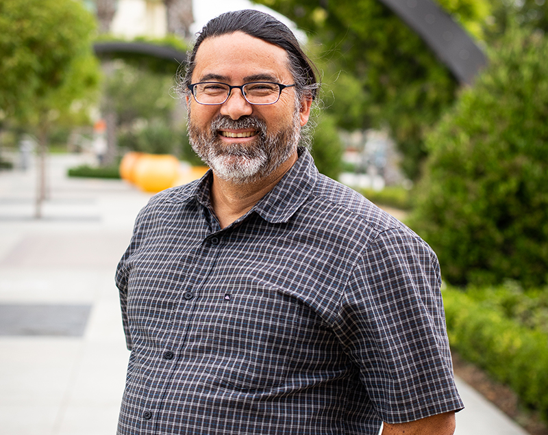 Daniel Nakaji wears a blue button down shirt with white stripes. He is outside on campus surrounded by greenery.