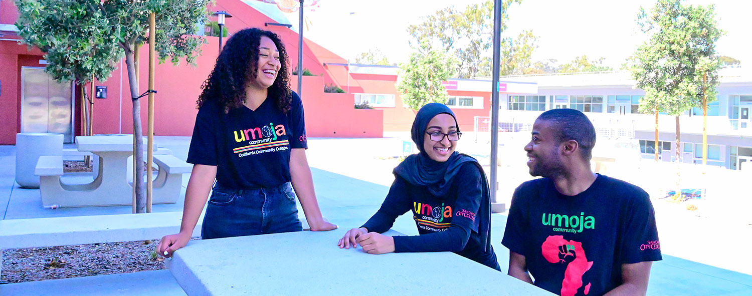 Three students sit at a cement table at the City College. Each student is wearing a black Umoja T shirt