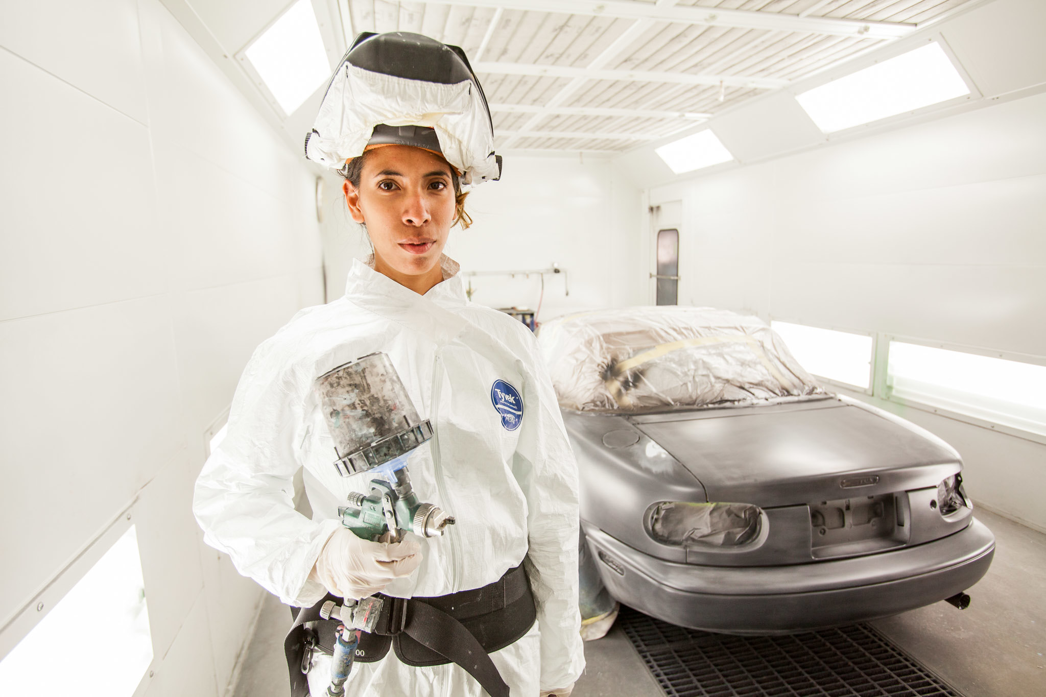 A student in the Auto Body and Paint Technician program wears a white jumpsuit and head covering while holding a paint gun. A silver car is in the background.