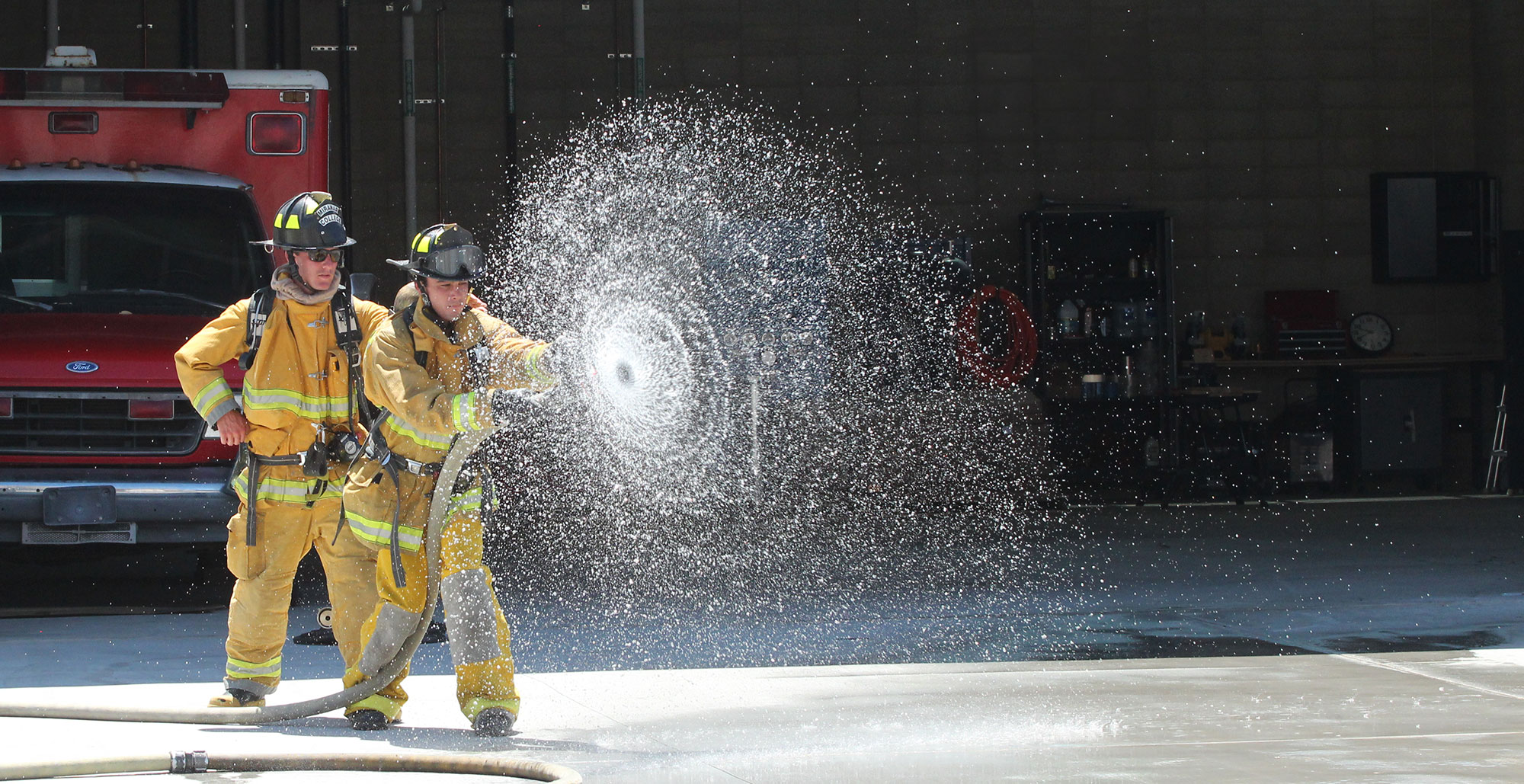 Two fire academy students in yellow fire gear use a fire hose. The water from the hose is making a spiral.
