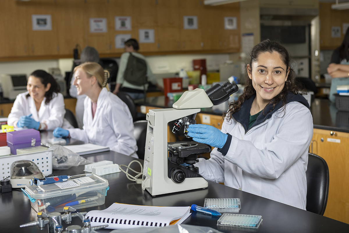 A student wearing a lab coat stands by a microscope in a science lab. Other students are in the background at other lab stations