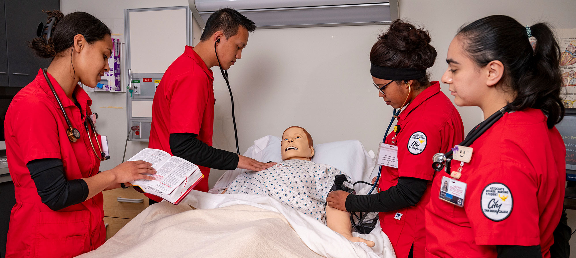 Four Nursing students in red scrubs surround a hospital bed. Two students are checking vital signs of a dummy that is in the bed