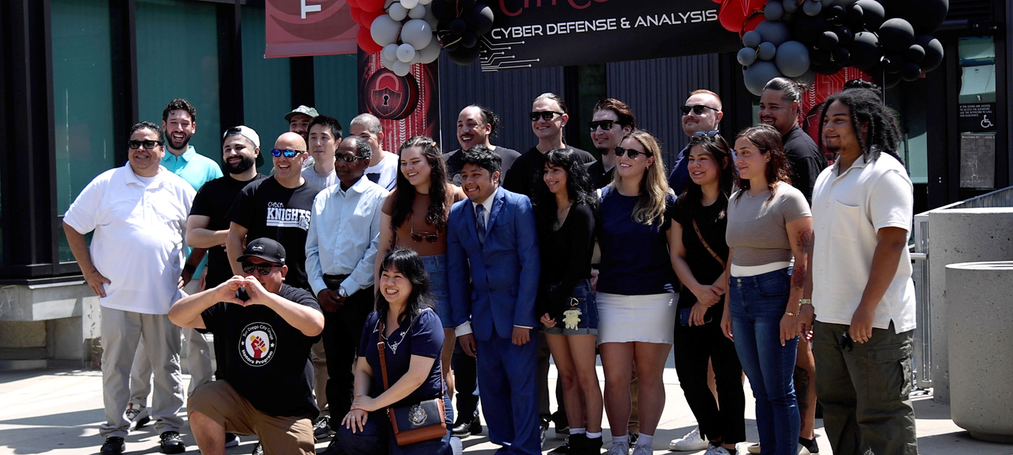 Twenty one people take a group photo outside a building at city college. There are red and black balloons and a city college banner behind them.