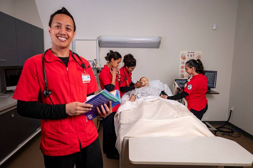 Four nursing students wearing red and black scrubs check on a dummy patient in a hospital bed