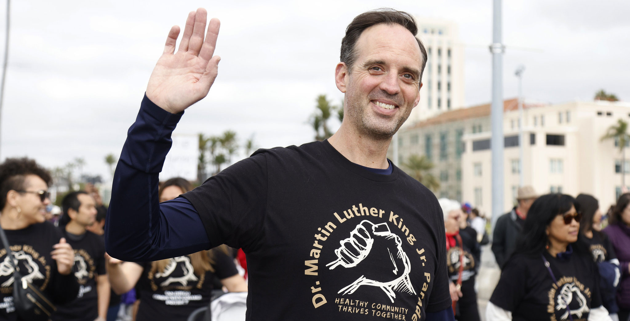 Greg Smith is wearing one of the black t-shirts distributed at the parade. There are two hands gripped together. Text reads MLK parade healthy community thrives together. Greg Smith is walking with the group and waving to the crowd. 