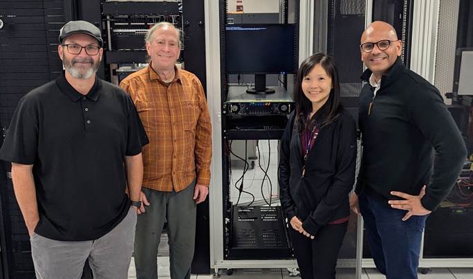 Four people stand in front of online servers and wires