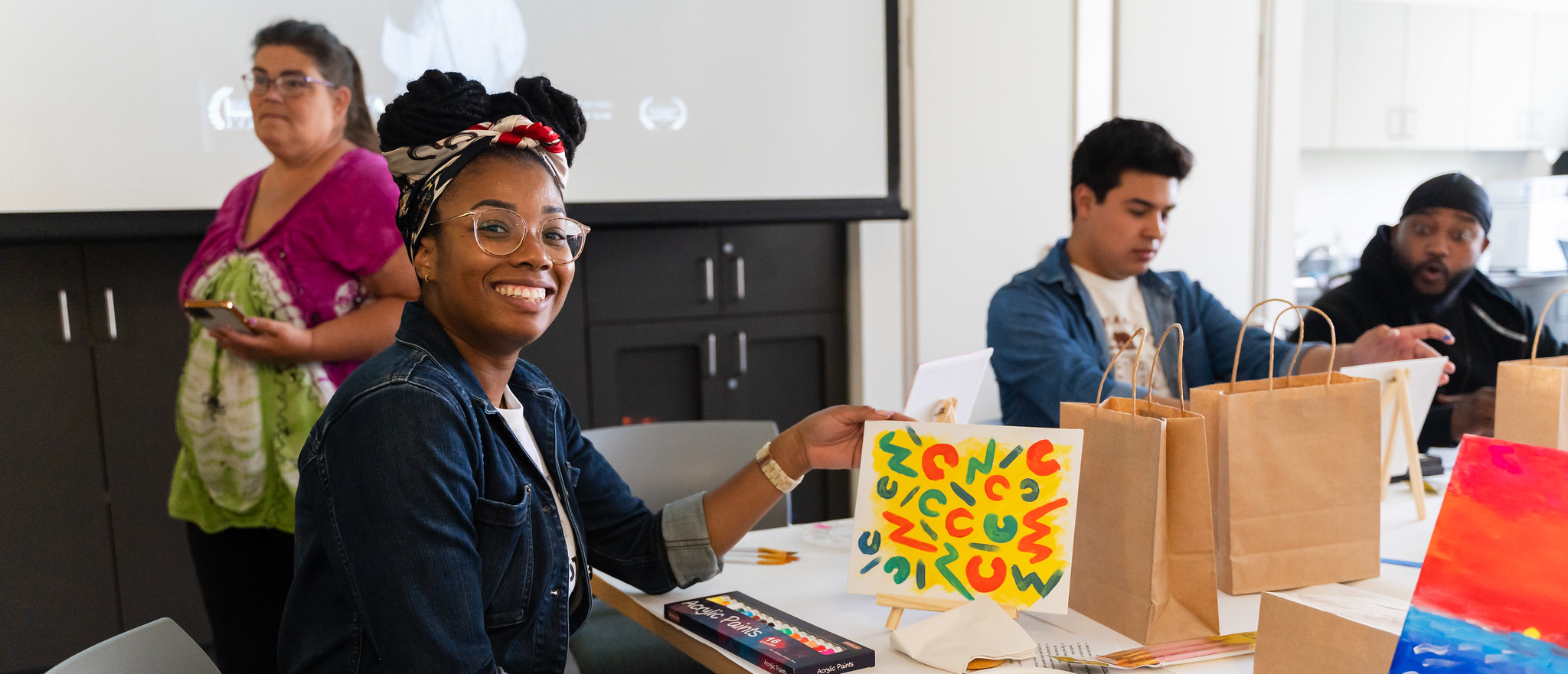 A student is sitting at a table and holding up a small painting that has a bright yellow background with red green and blue squiggles