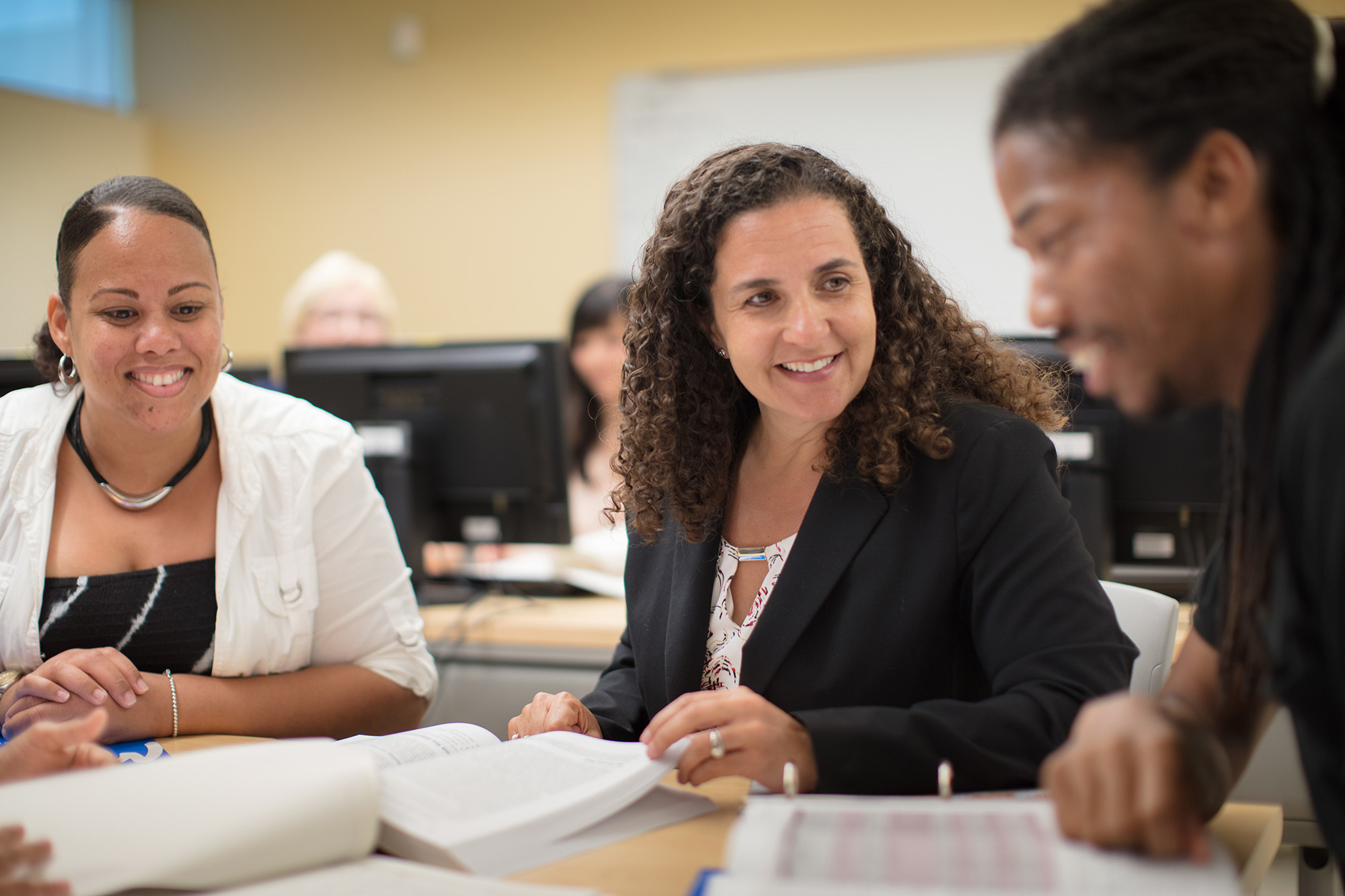 Professor Connie Renda wearing a black suit at a table talking to two students. There are books and notebooks on the table.