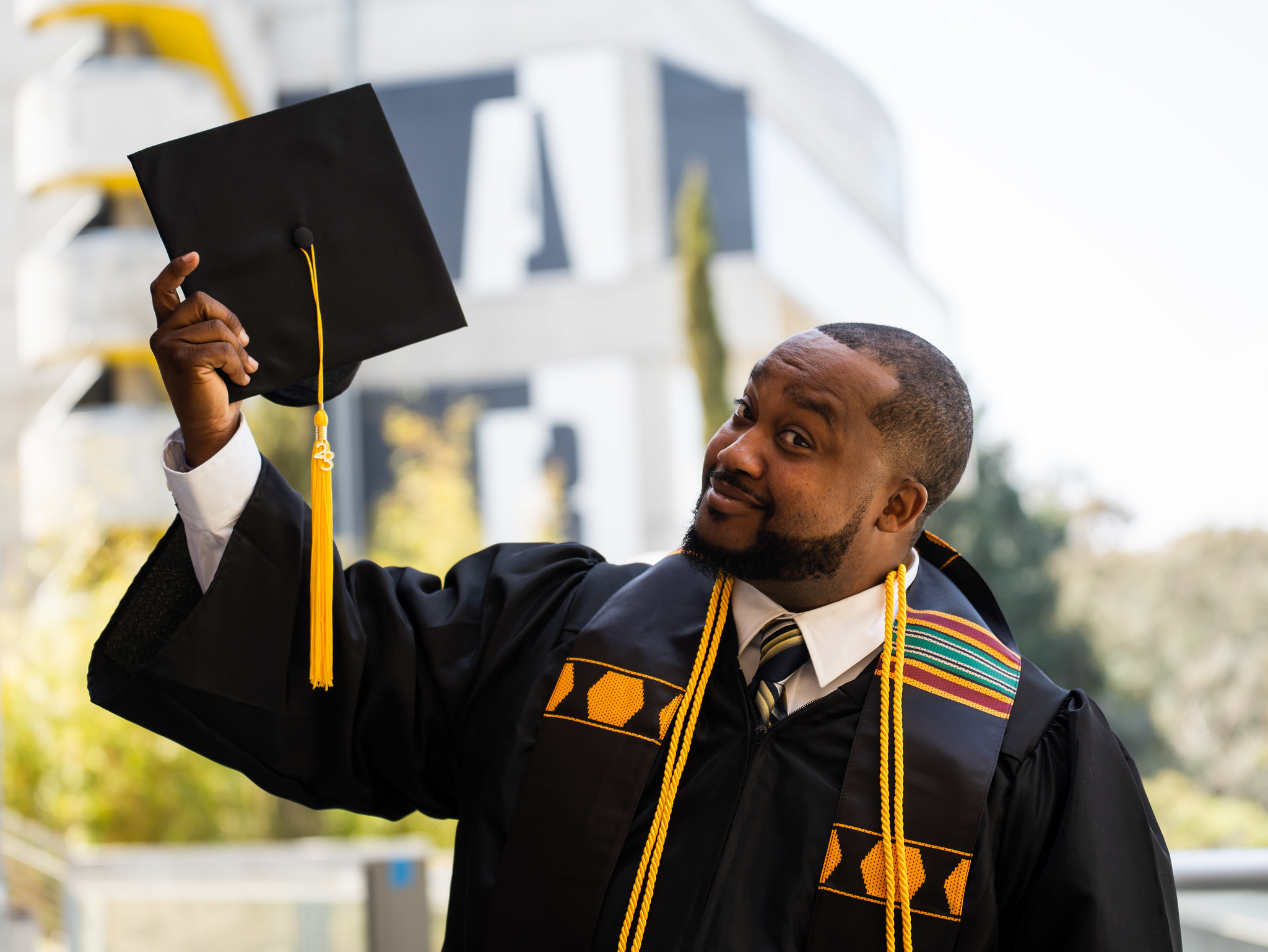 A graduate in a black graduation gown with a colorful stole and gold chords around his neck holds up his graduation cap with a gold tassel