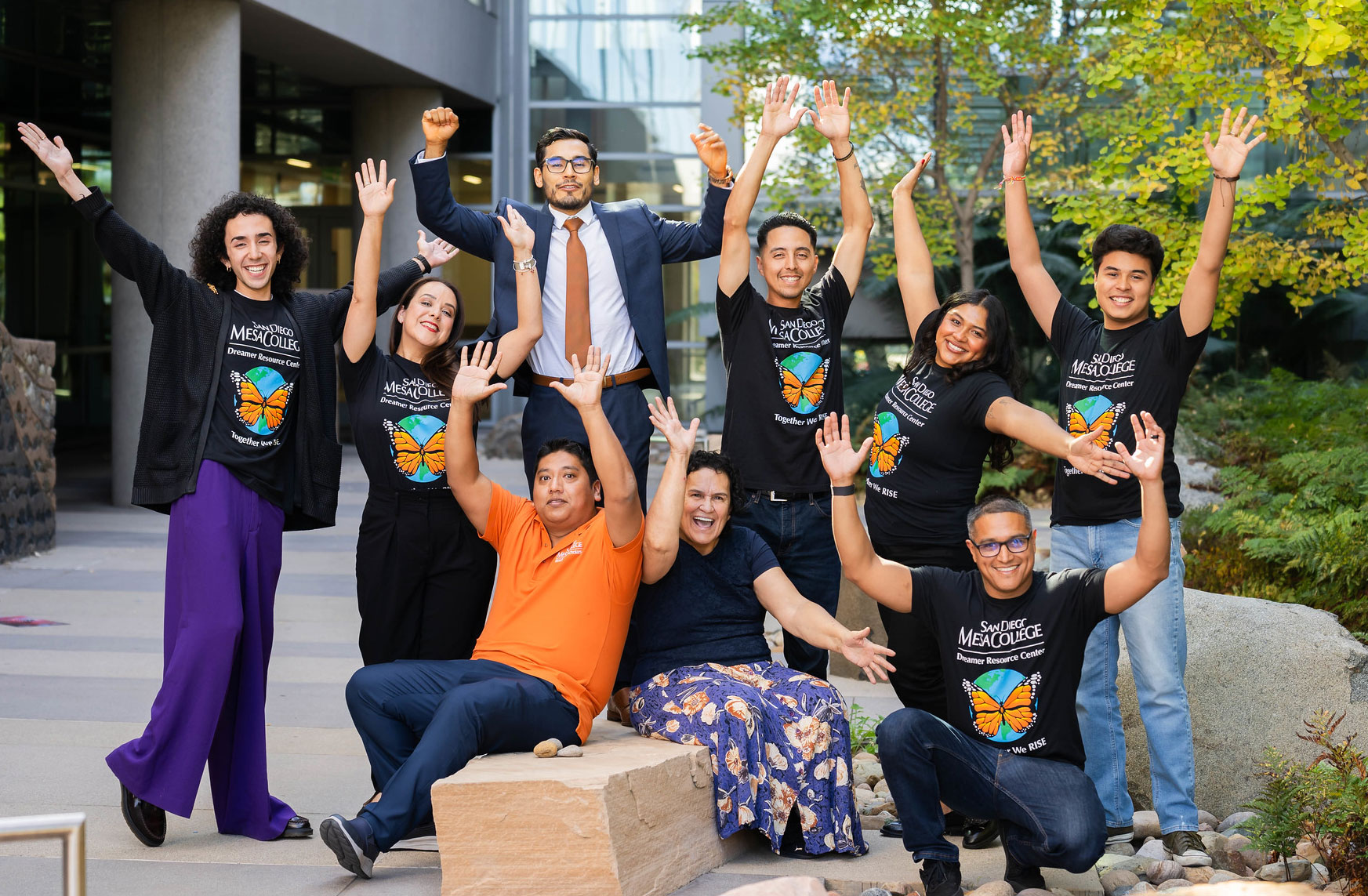 Nine people are raising their hands cheering outside in a courtyard of a campus builing. five of them are wearing black tee shirts with butterflies on them representing undocumented students 