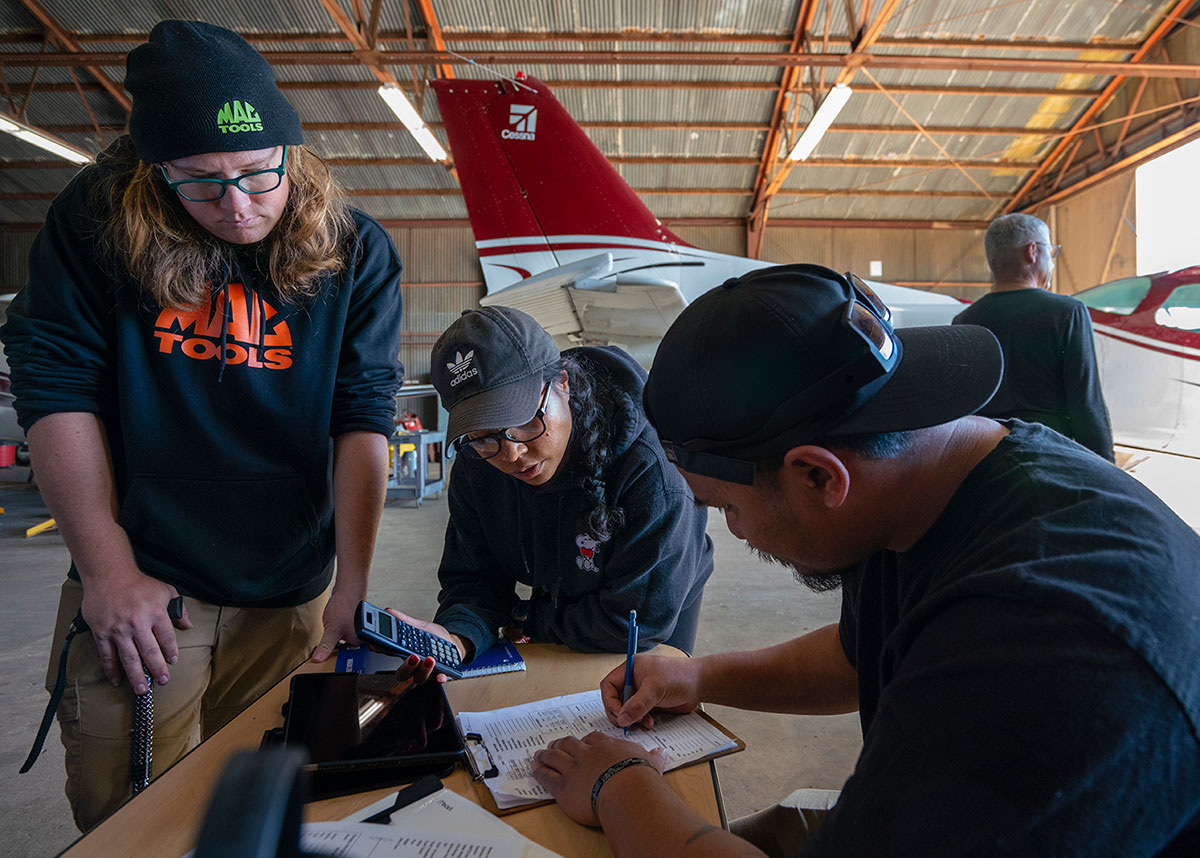three students work on calculations at a table in a hangar. A small plane is in the background.