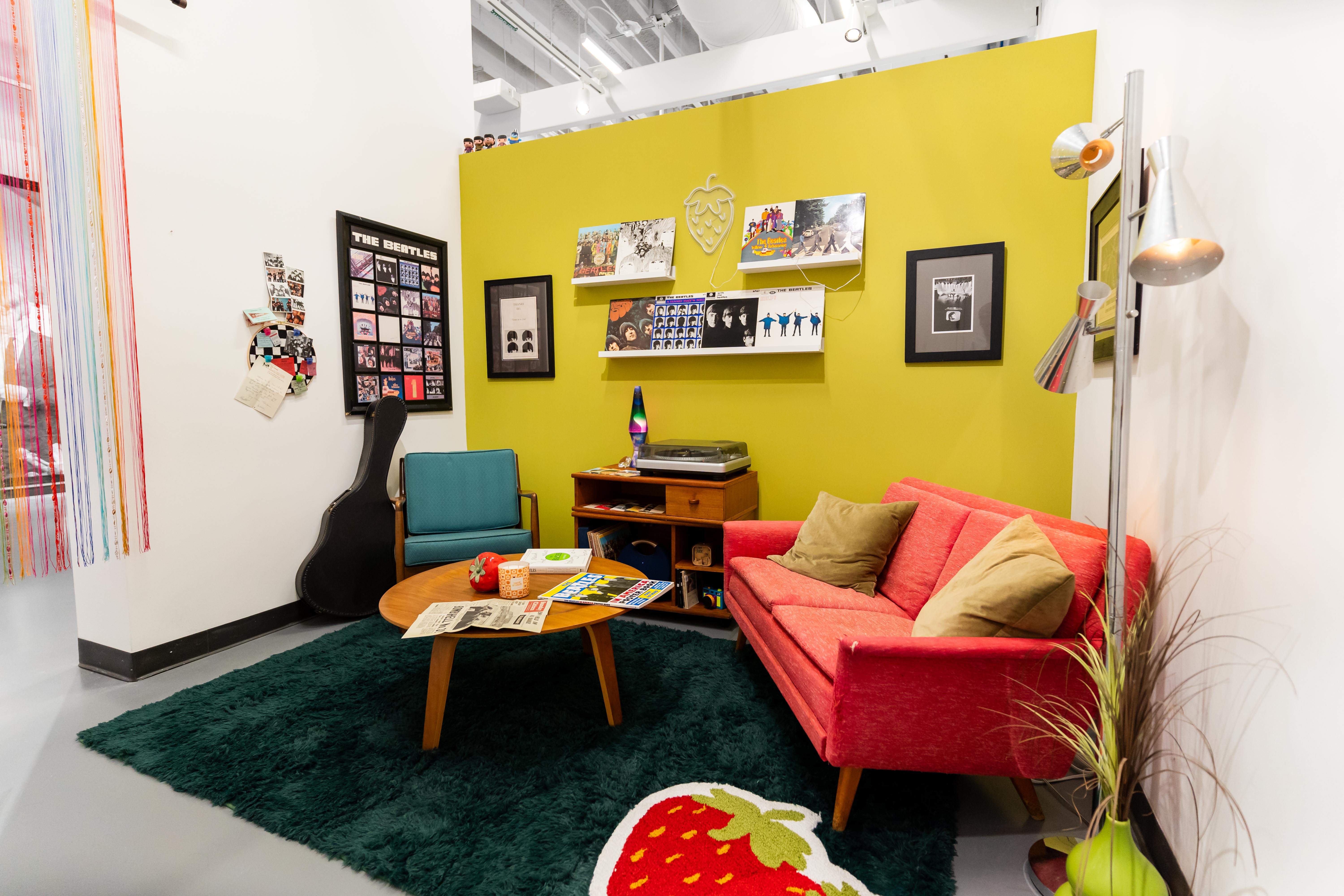 A corner of the gallery set up like a room with a bright orange chair a guitar leaning on a wall and Beatles pictures hanging on the wall.