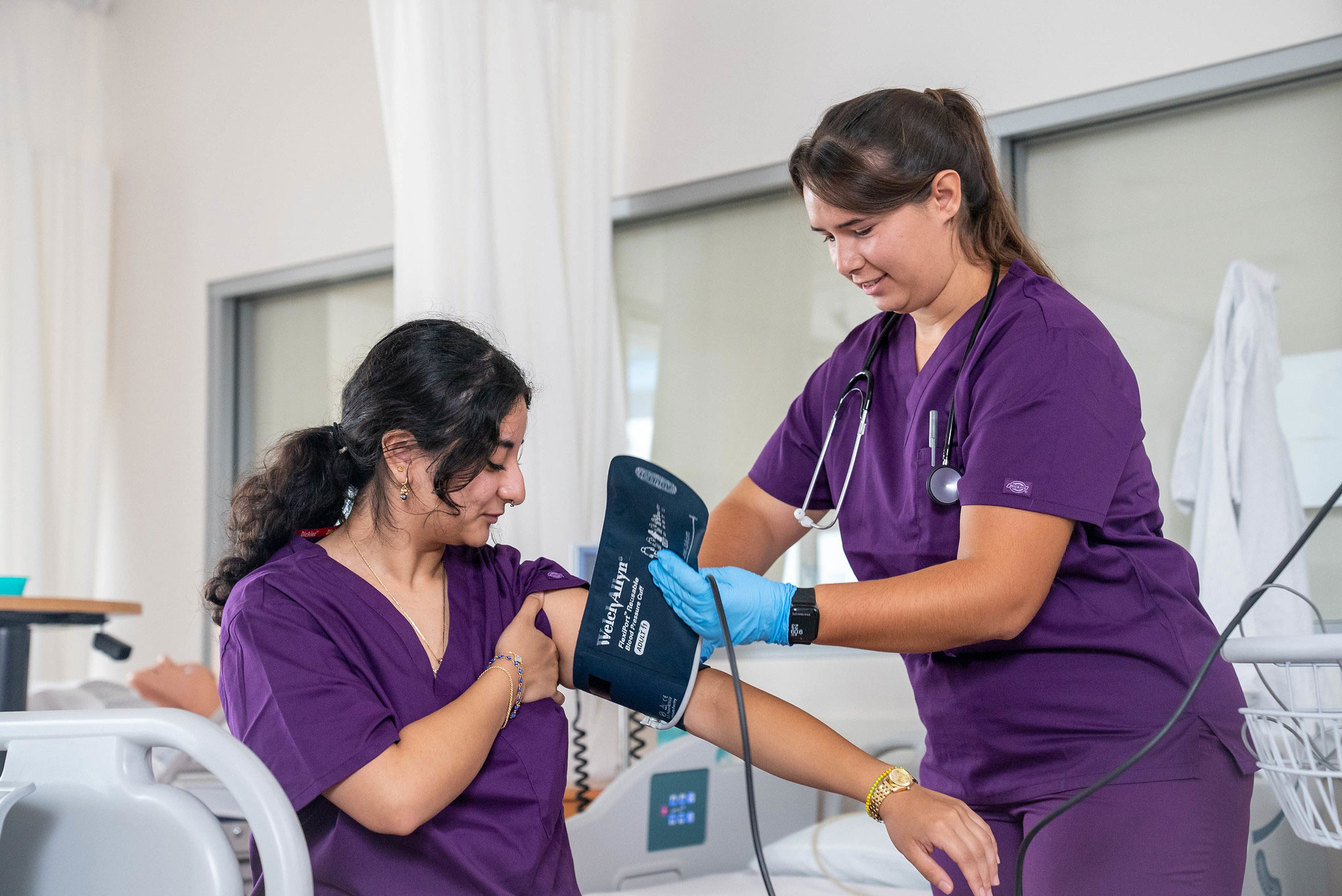 Two students in purple scrubs. One is putting a blood pressure cuff on the other's arm.