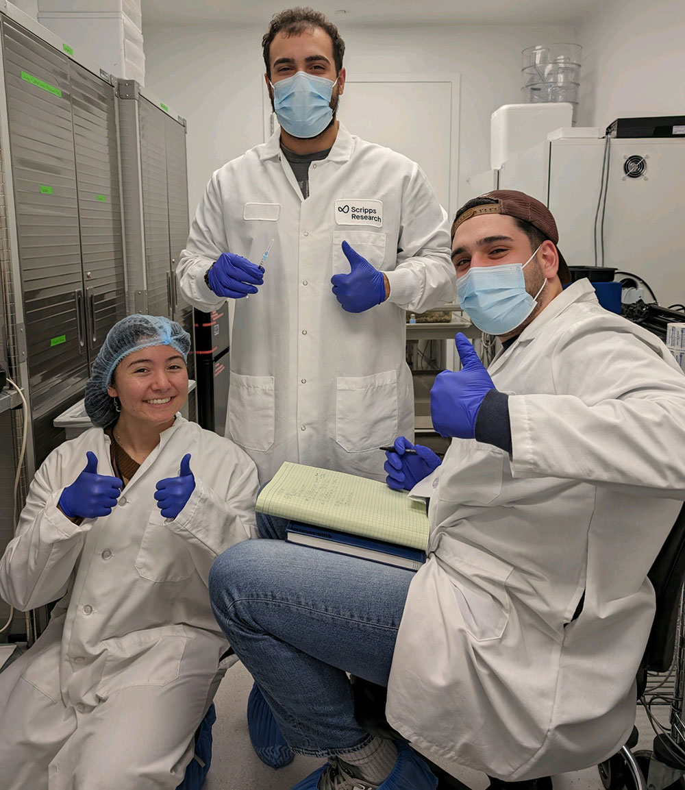 Three interns in lab coats, purple latex gloves and facemasks give double thumbs up