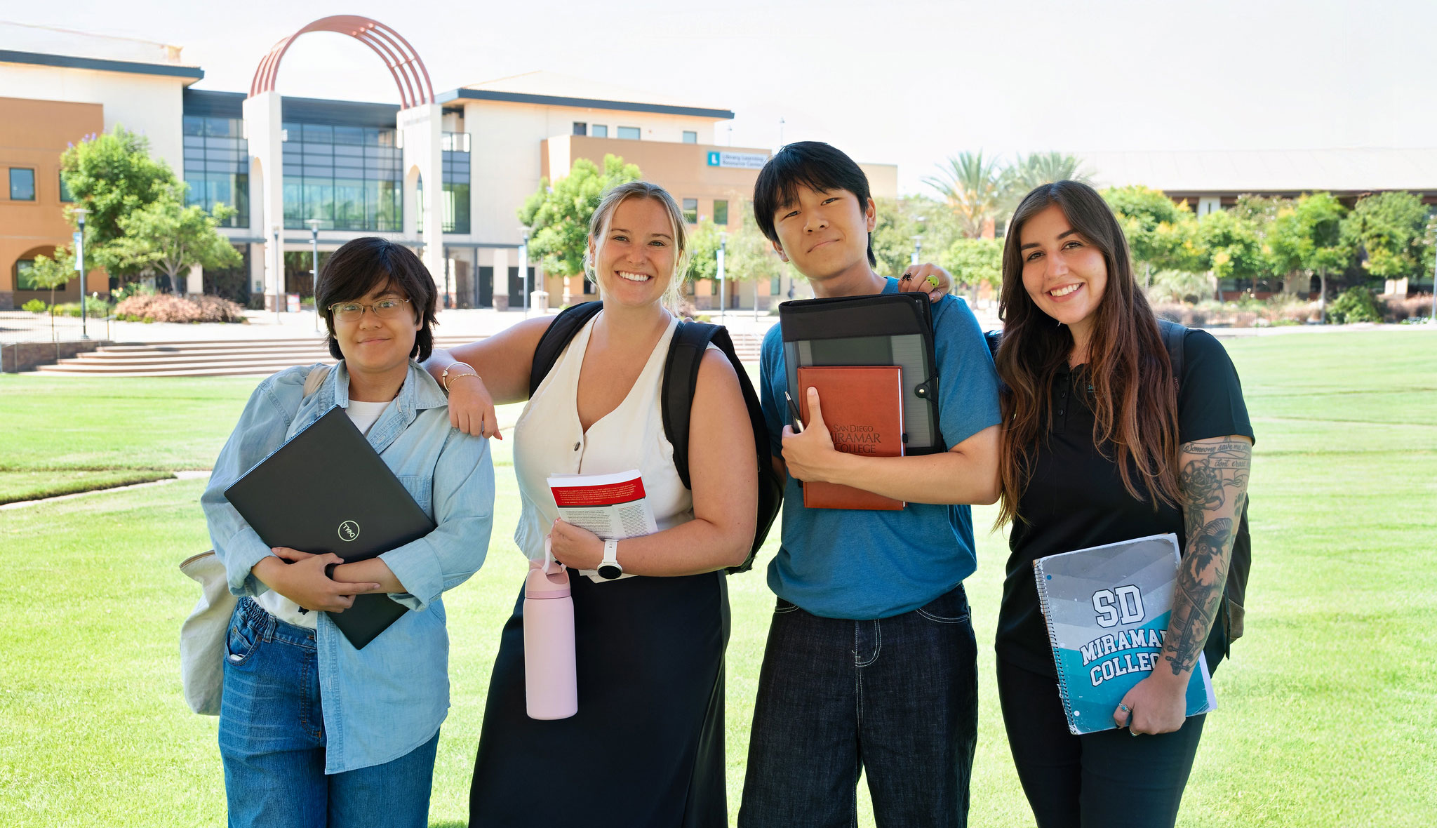 Four Miramar College students holding books and carrying backpacks on the grass in front of the Administration Building on campus.