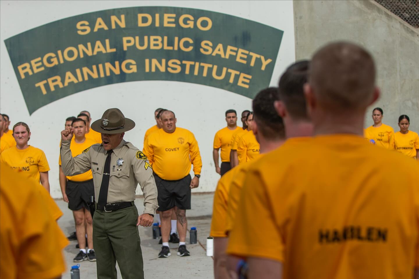 An instructor in a brown officer's uniform speaks to cadets in orange shirts lined up.