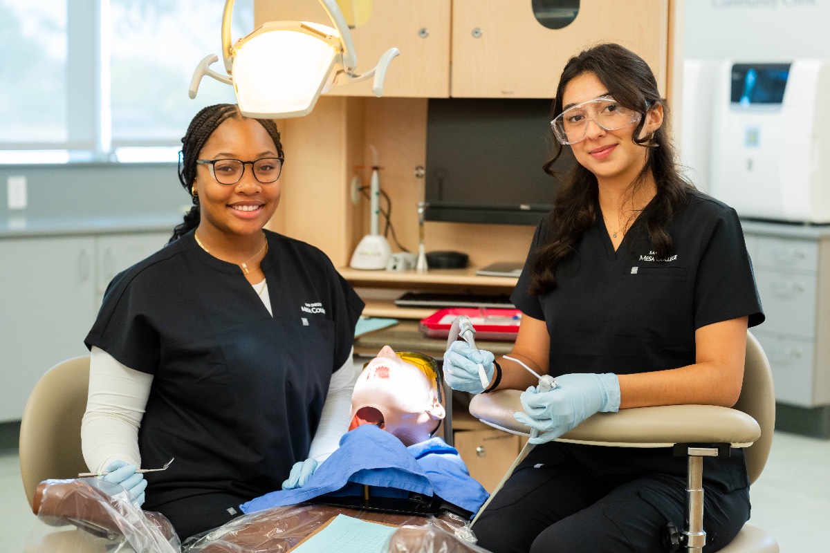 Two students wearing black medical scrubs sitting by a dental chair with a dummy in the chair