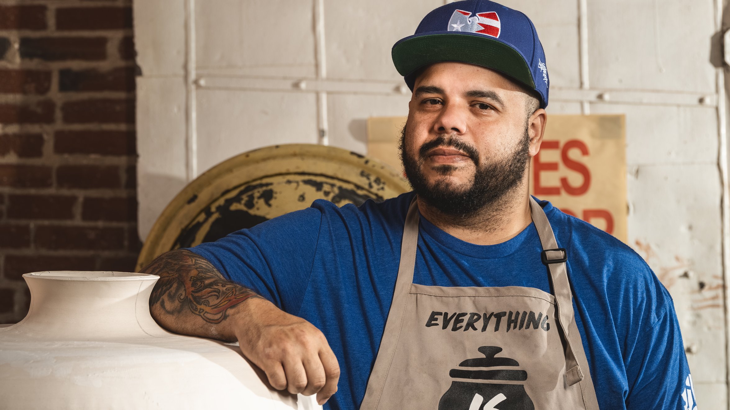 Roberto Lugo in a studio wearing a blue baseball cap and a tan apron leaning on a large pottery vase