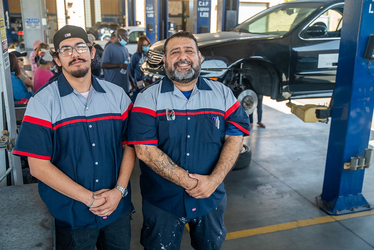 Daniel and Luis wearing blue mechanic pants and shirts. A car is on a lift behind them.