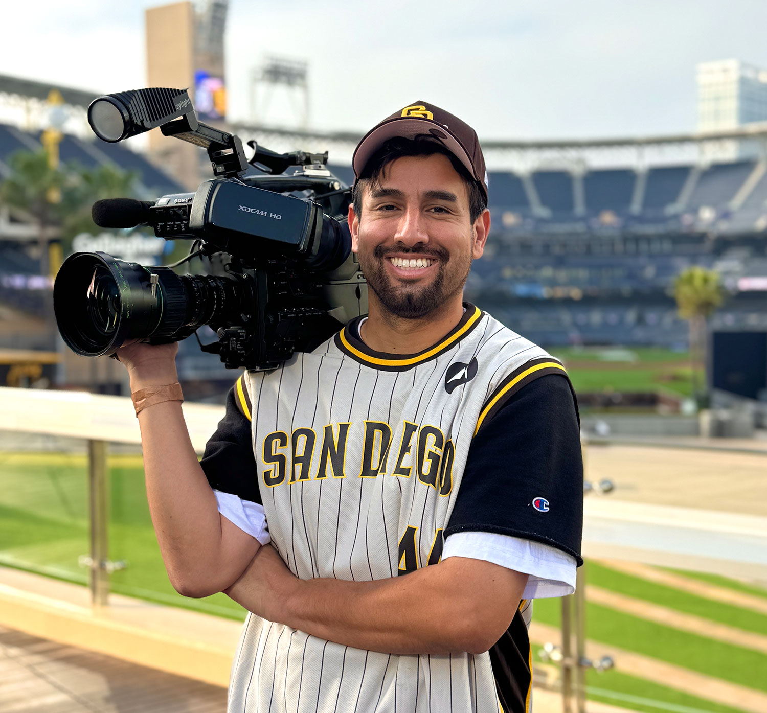 David Pradel holding a professional video camera on his shoulder wearing a Padres hat and jersey with Petco Park in the background.