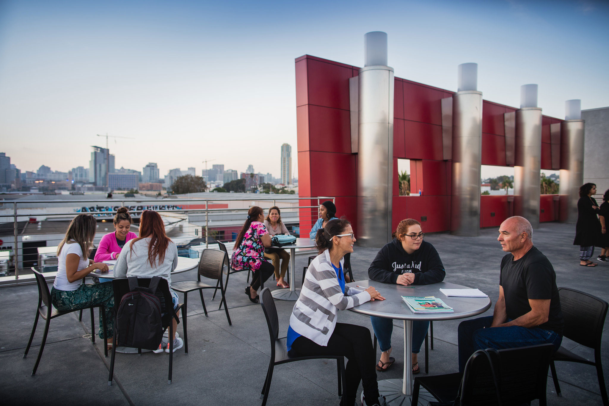 Nine people sit at tables on the rooftop of the campus in Barrio Logan. The San Diego skyline is in the background. 