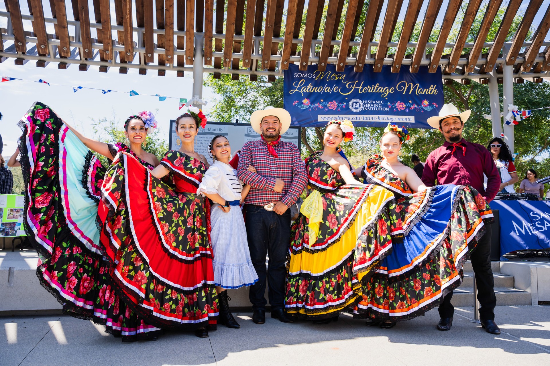 Five women in colorful dresses and a two men dressed as cowboys perform dances at a hispanic heritage celebration.