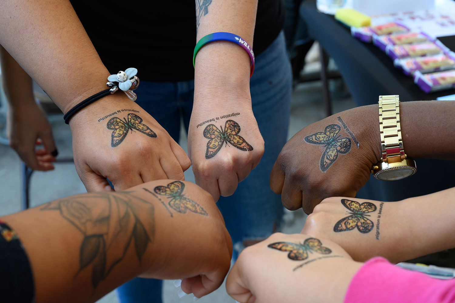 Six people with their hands together in a circle all have a butterfly tattoo on the back of their hand
