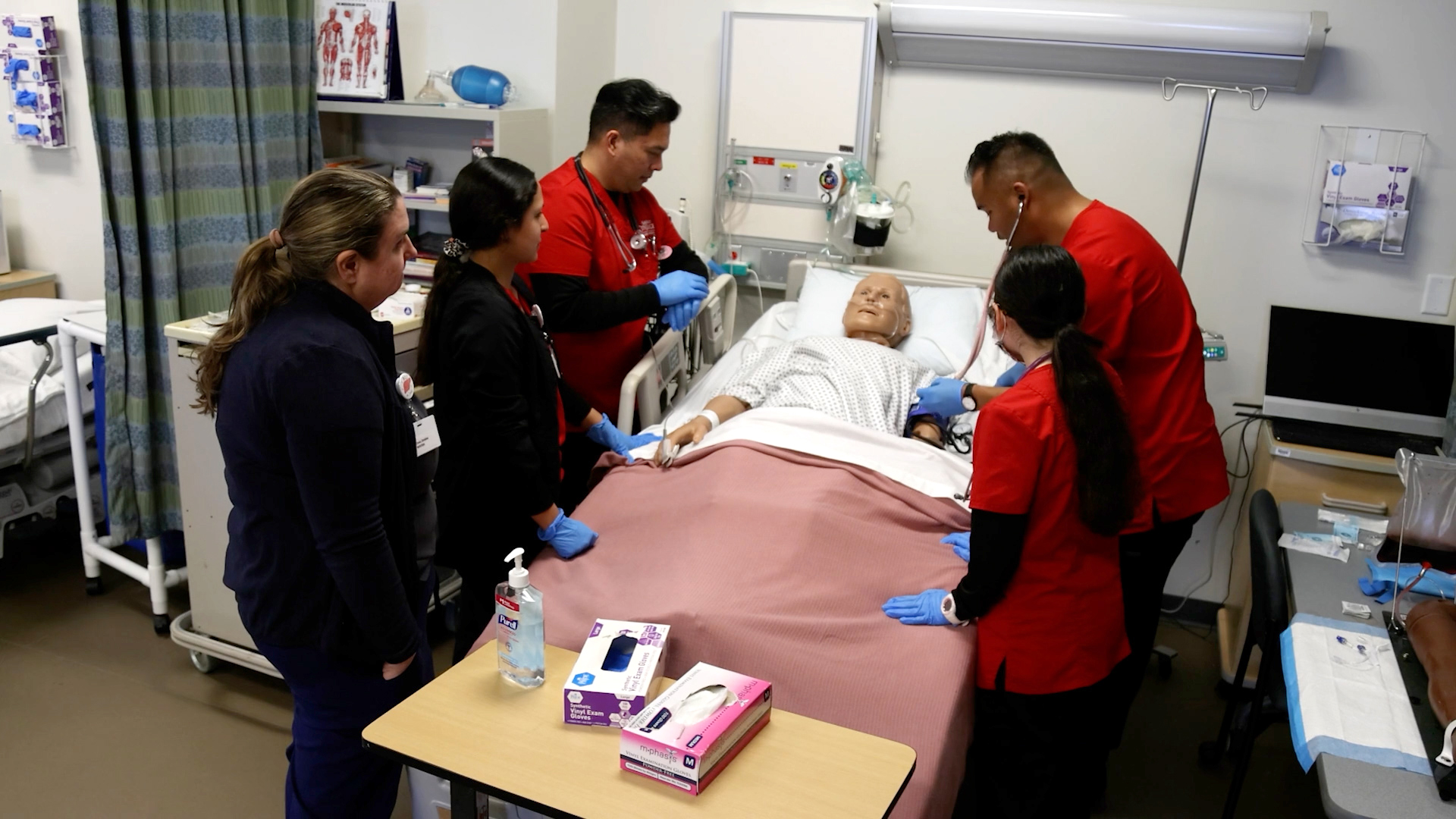 Five students in red medical scrubs around a hospital bed with a dummy patient