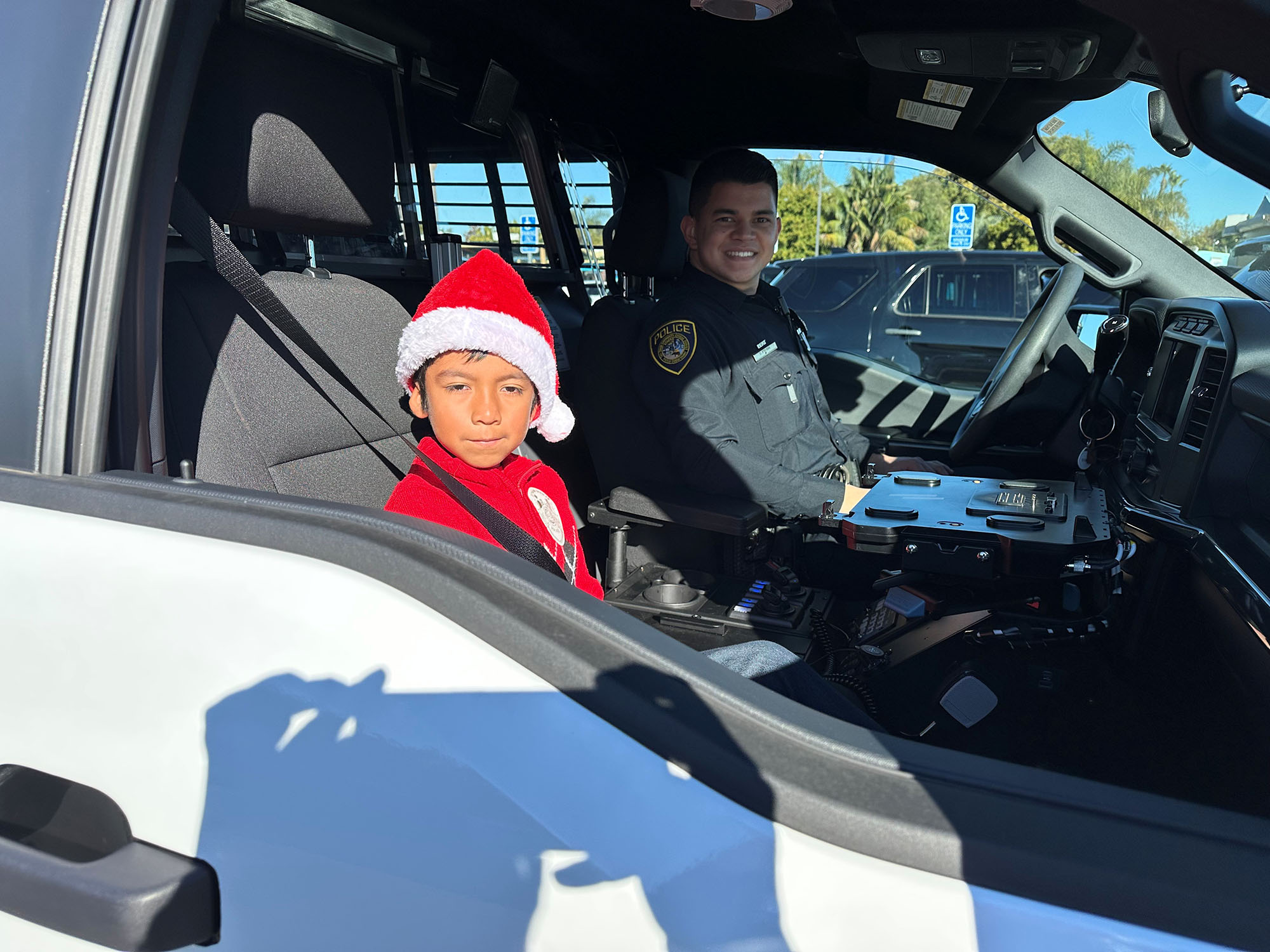A child wearing a Santa hat sits in the front seat of a police SUV. An officer is driving.