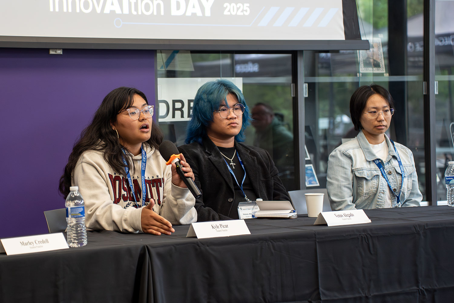 
Three people on a panel sitting at a table. One student is speaking to the audience.
