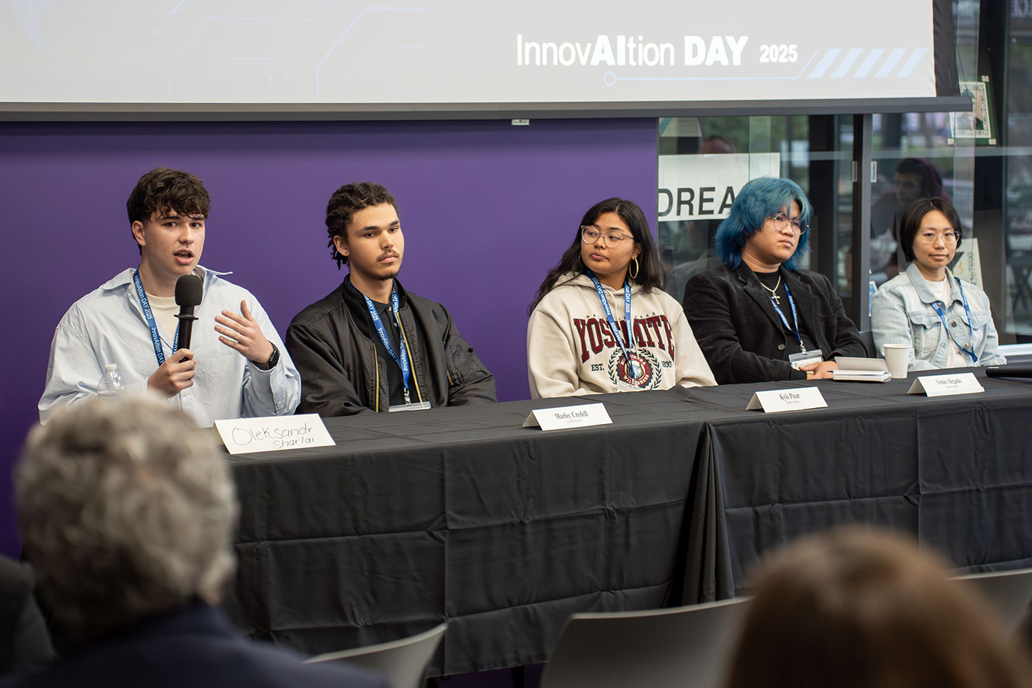 
Five students on a panel sit a table. One is speaking into a microphone.

