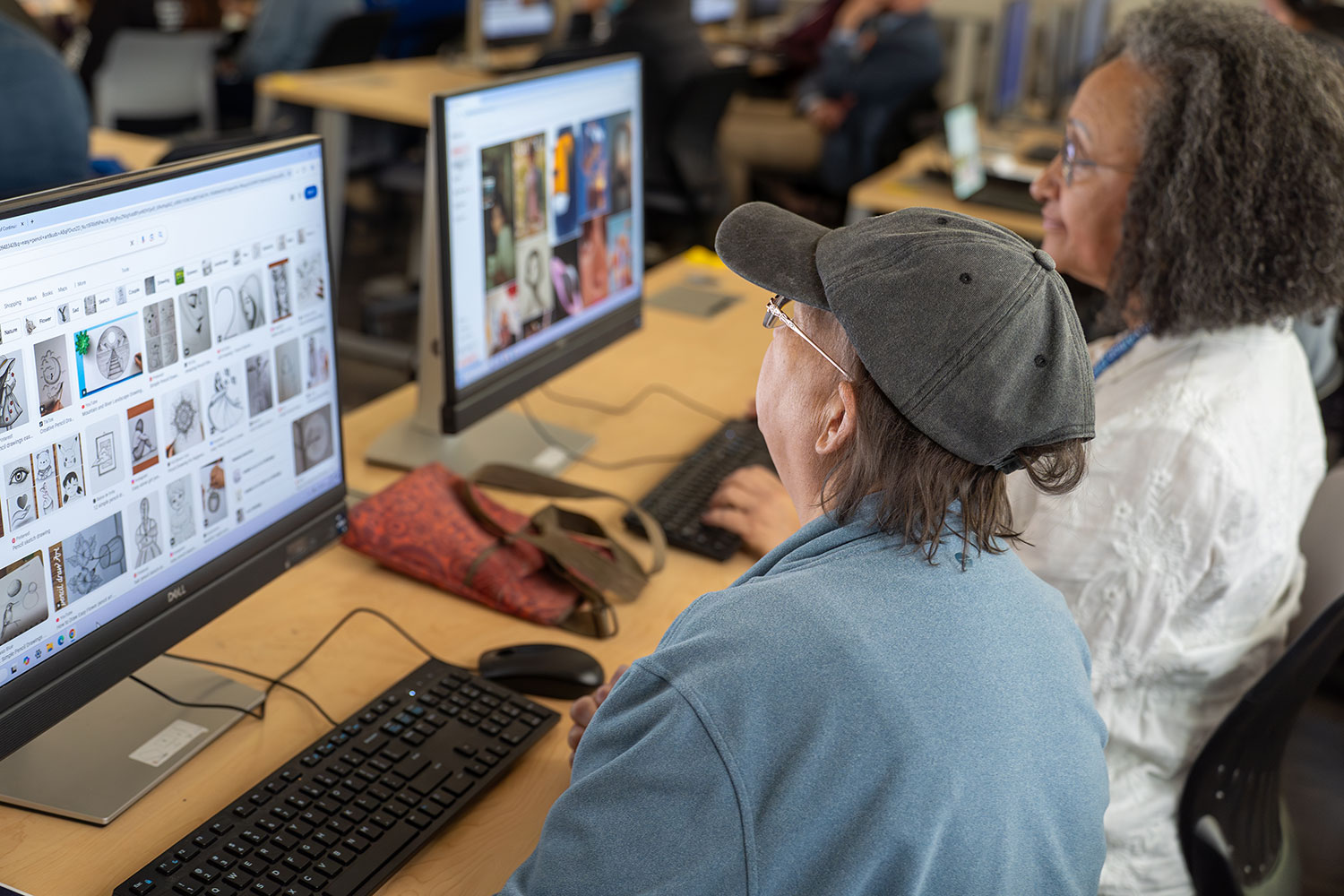 
Two women looking at a desktop computer screen in a classroom of computer stations.
