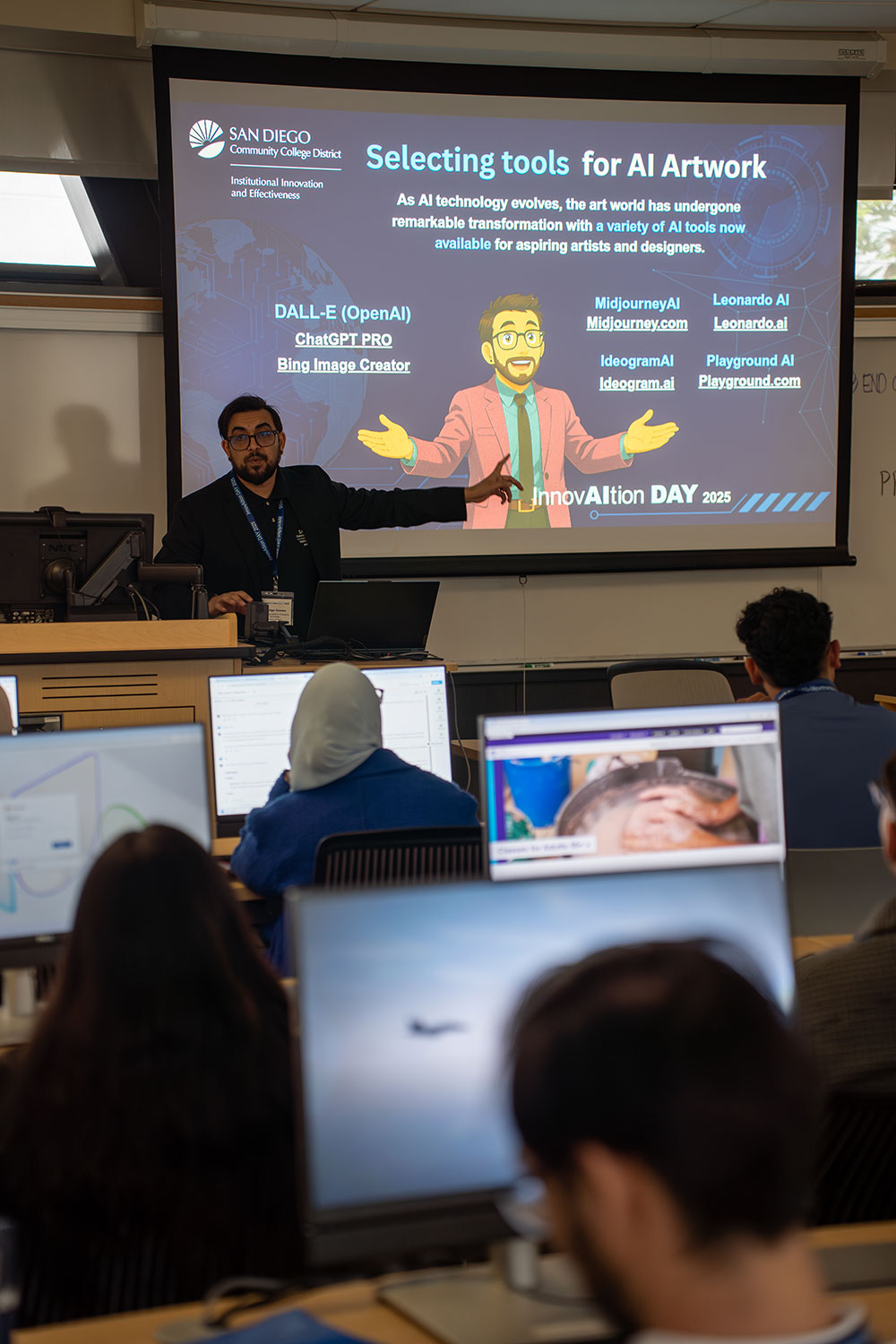 
People sitting at computer stations listening to a speaker pointing to a slide on a screen.
