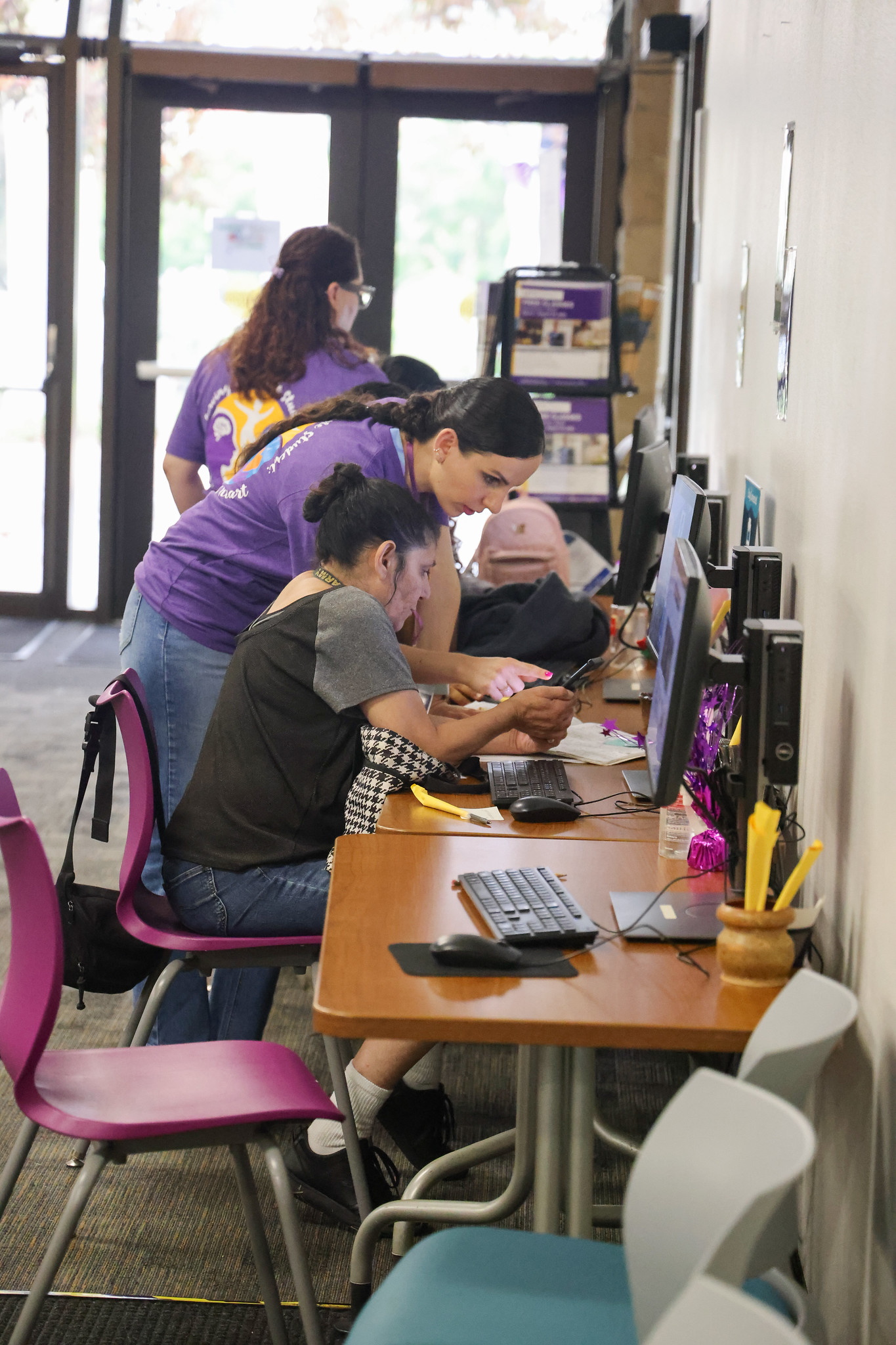 
A worker leans over to help a student sitting at a table and working on a computer.
