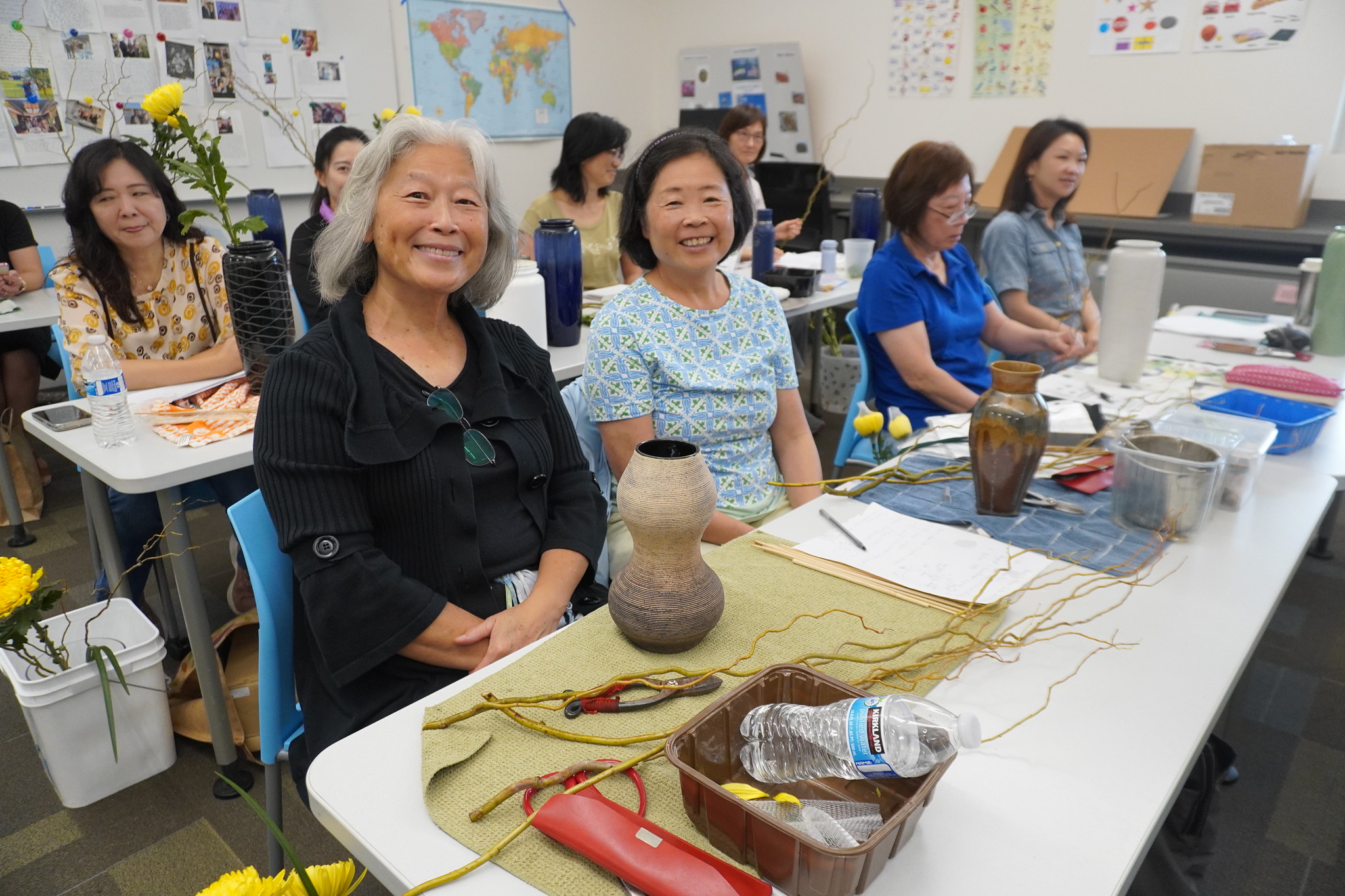 
Students sit at tables in front of vases in a flower arranging class.
