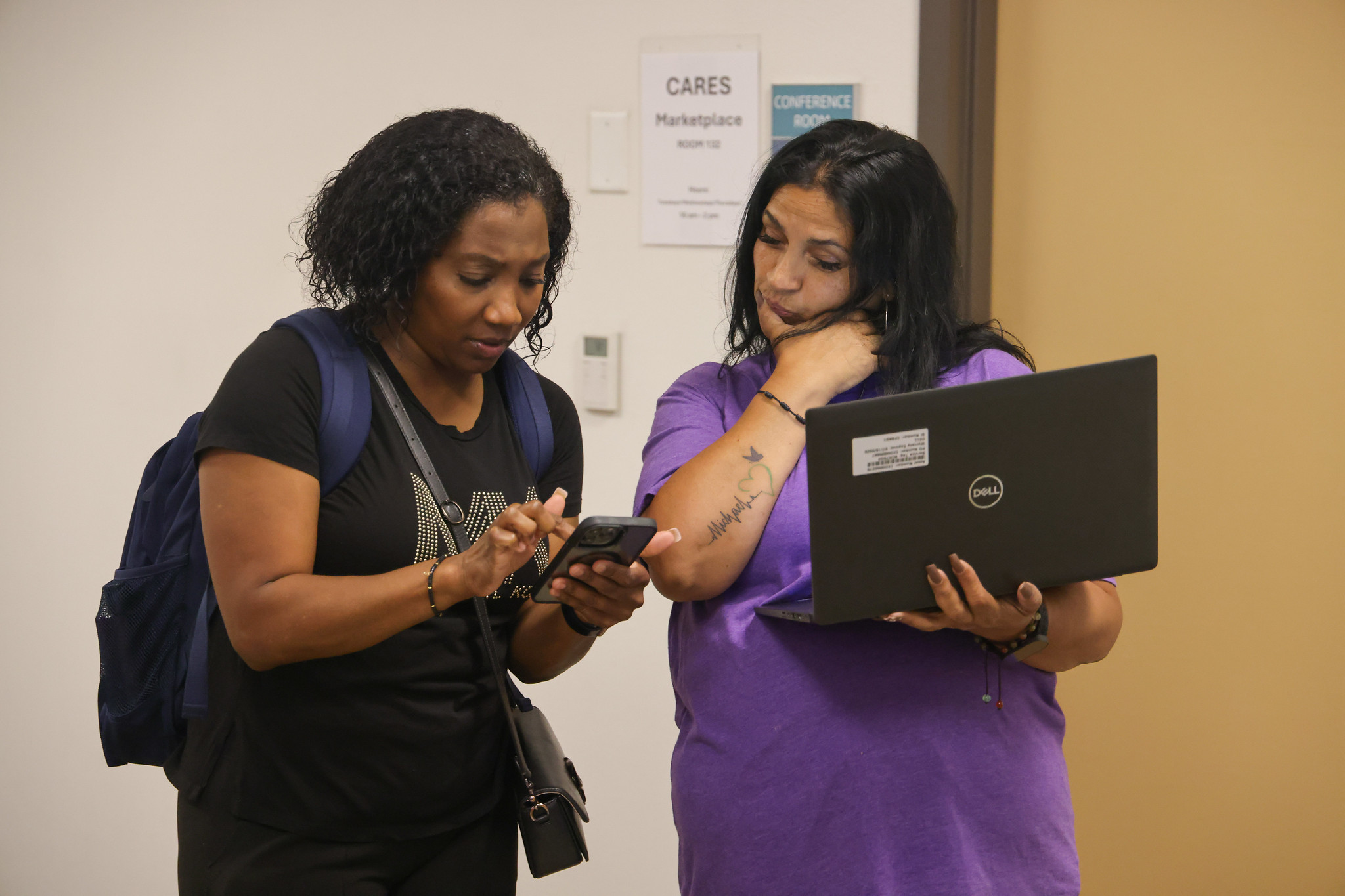 
A student is looking something up on her phone while an employee holding an open laptop looks on. 
