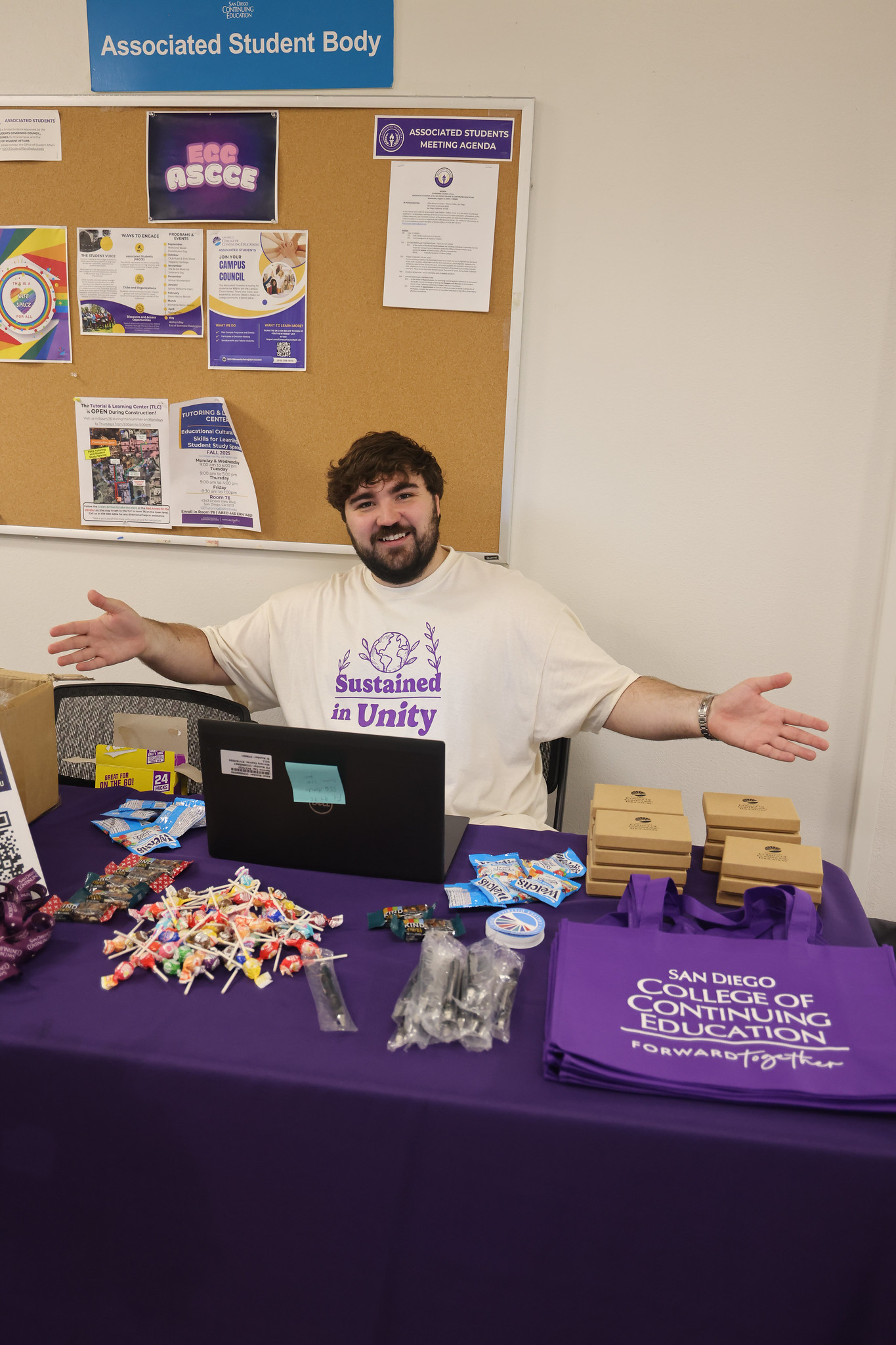 
A worker at a welcome table opens his arms toward the table to showcase candies, tote bags and other swag that is being given away to students.
