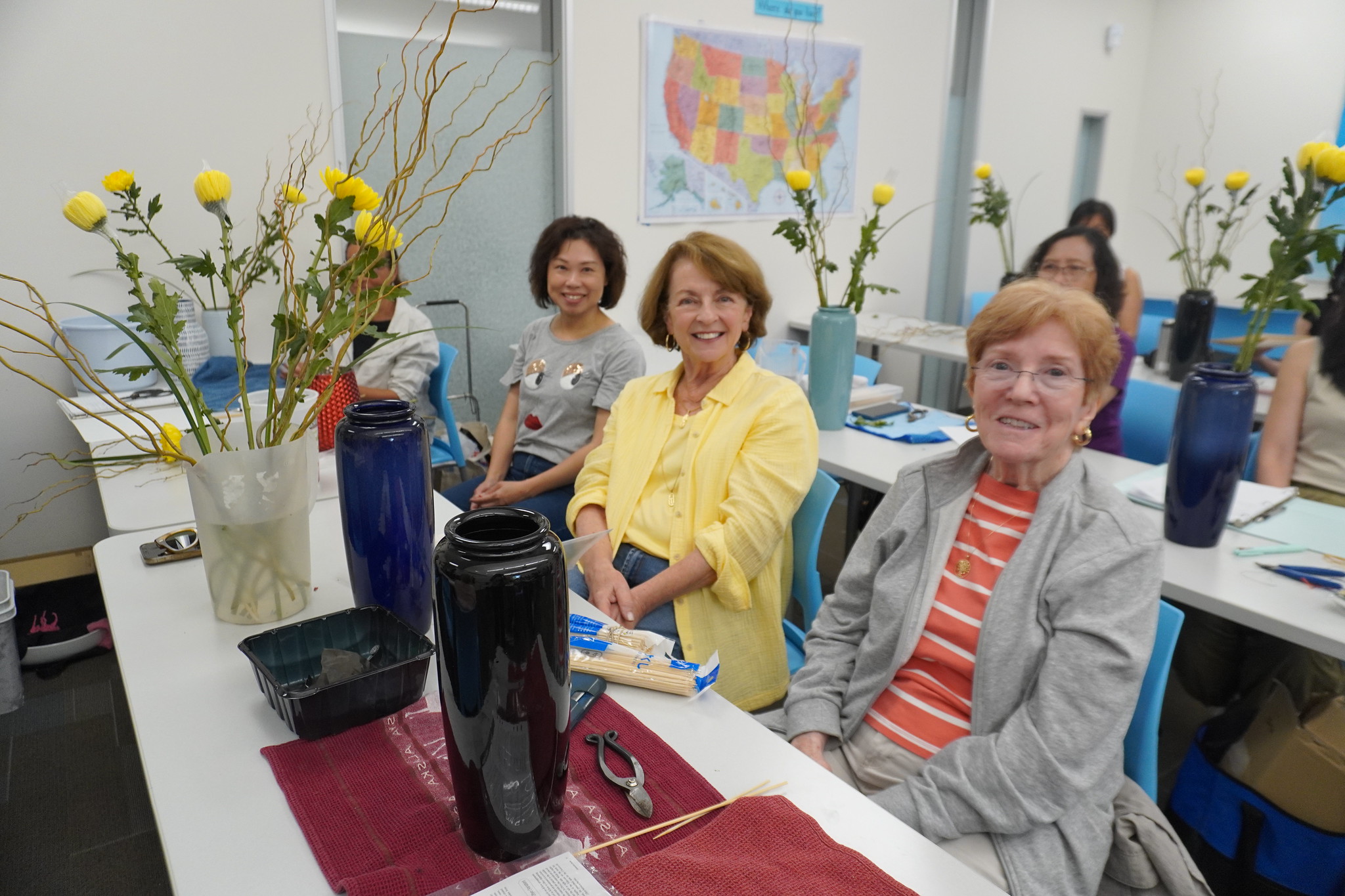 
Students sitting at tables with vases and flowers in front of them for a flower arranging class. 
